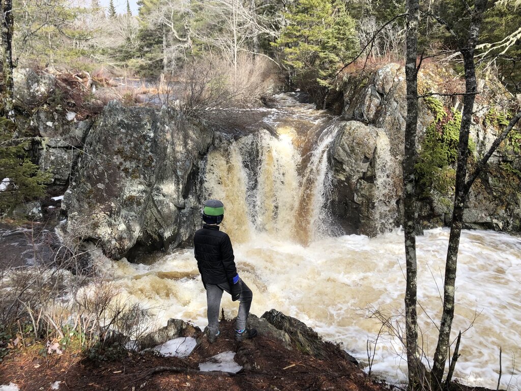 A child wearing a black jacket and green hat stands on a rocky ledge looking at a small waterfall cascading between large rocks in a forest near Halifax. Fast moving, foamy water rushes below, with evergreen trees and patches of snow surrounding the scene, highlighting a rugged outdoor spot for exploring waterfalls near Halifax.