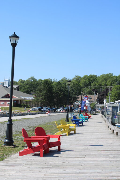 A row of brightly colored Adirondack chairs—red, yellow, blue, and green—lined up along a wooden boardwalk in Pictou, Nova Scotia, with lampposts and a view of trees, buildings, and parked cars under a clear blue sky.