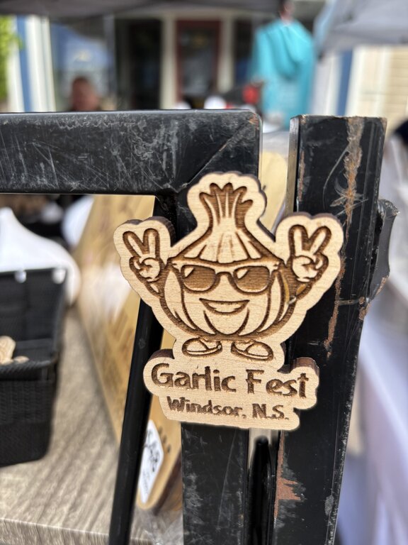 A wooden magnet or ornament shaped like a smiling garlic bulb wearing sunglasses and flashing peace signs, engraved with the text “Garlic Fest Windsor, N.S.” It’s displayed on a black metal stand at a festival booth.