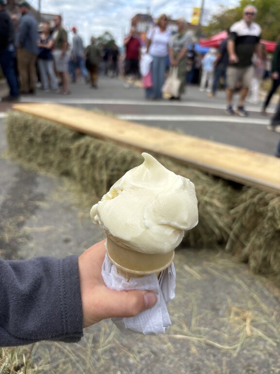 A child’s hand holding a vanilla (actually garlic flavoured) ice cream cone wrapped in a napkin, with a blurry background showing hay bales and a crowd of people at an outdoor festival.