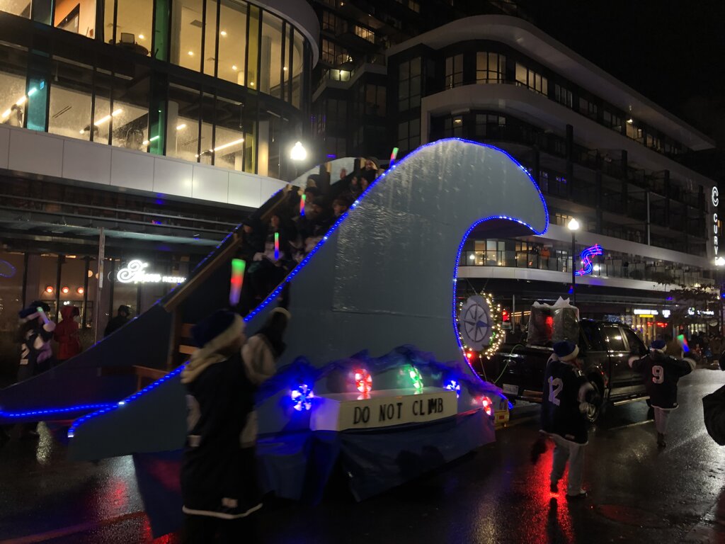 A festive parade float shaped like a giant ocean wave with glowing blue lights carries children waving light sticks, while people in matching outfits walk alongside on a wet city street at night. A sign on the float reads "DO NOT CLIMB" and a truck tows the display past a modern glass building.