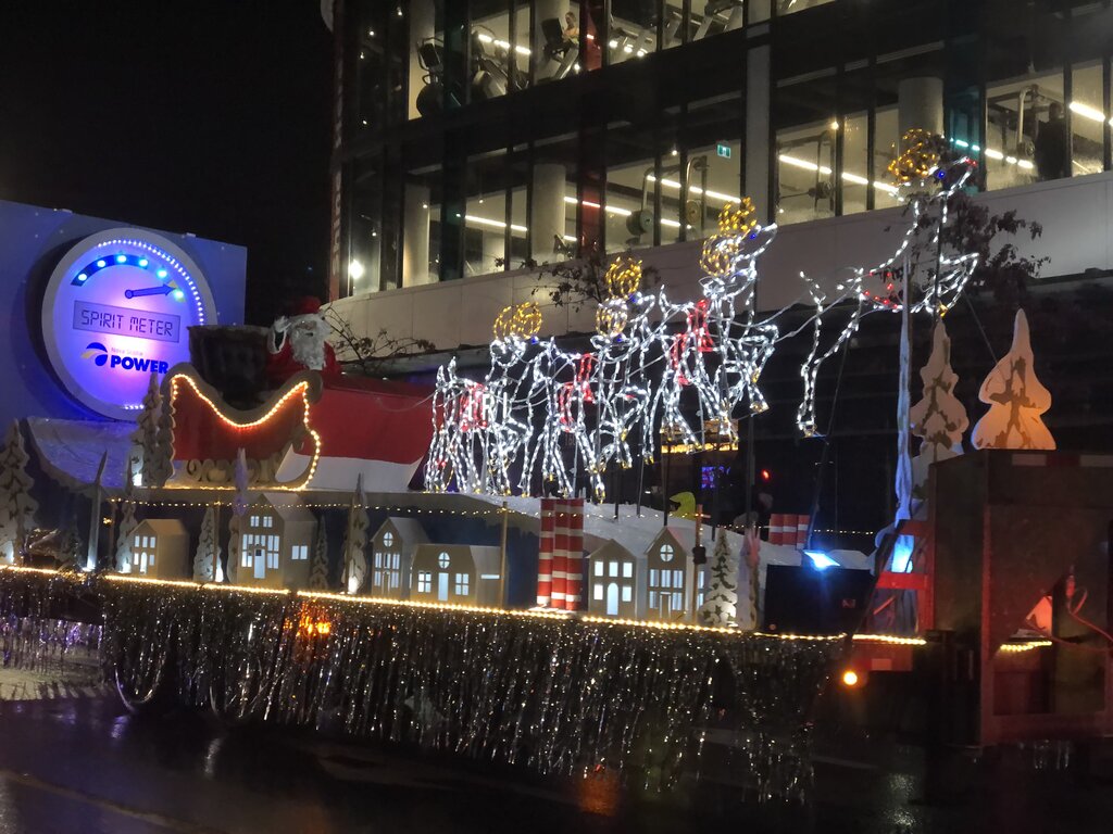 A brightly lit Christmas parade float features Santa Claus in a red sleigh led by sparkling reindeer made of white lights, flying over a miniature illuminated village. The float includes tinsel garlands, stylized pine trees, and a giant “Spirit Meter” sign in the background, creating a cheerful holiday scene.
