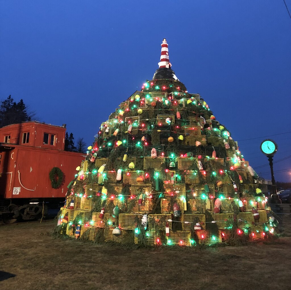 A large Christmas tree structure made entirely of stacked green lobster traps, decorated with buoys and colorful holiday lights, topped with a mini red-and-white lighthouse; set beside a red caboose in Musquodoboit Harbour