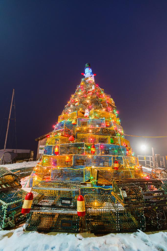 square lobster traps are stacked to look like the shape of a christmas tree. buoys and lights decorate the tree and there is a snowman on top