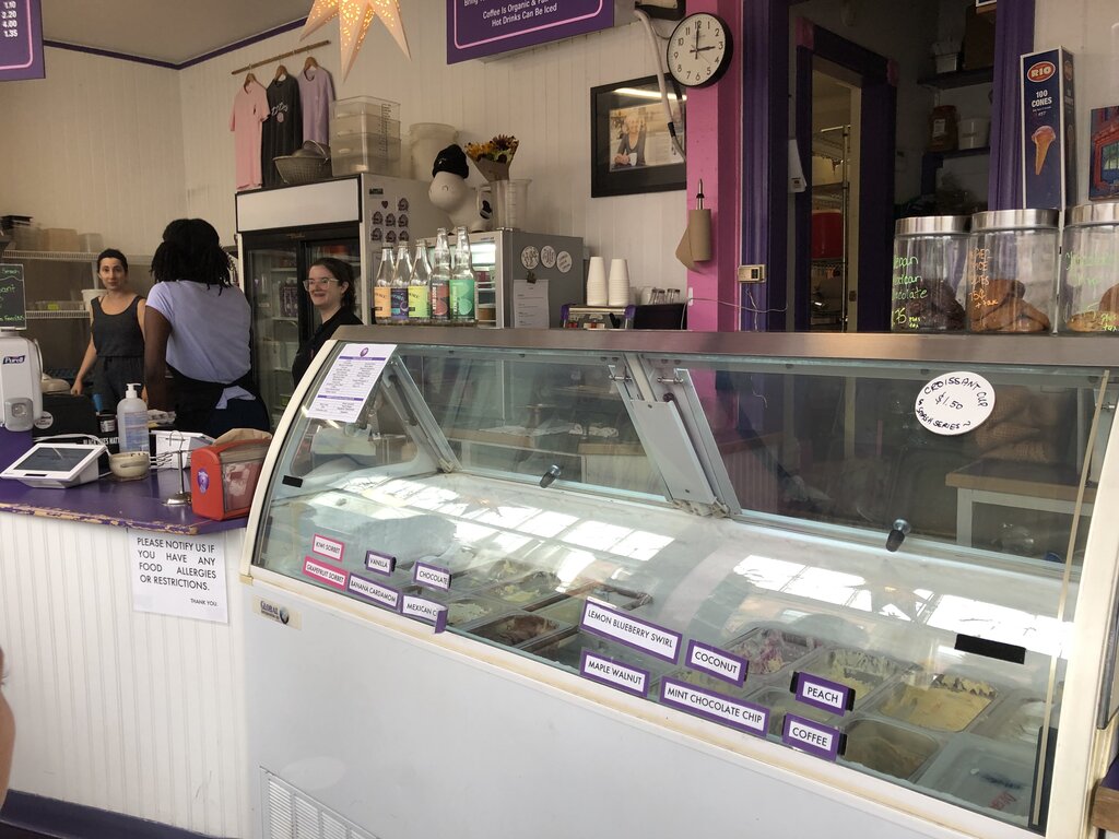 Inside Dee Dee’s Ice Cream shop showing a glass ice cream counter with labeled flavors like Maple Walnut, Coconut, and Coffee. Three staff members are behind the counter smiling, with colorful soda bottles and T-shirts on display in the background. A sign on the wall reminds customers to notify staff about food allergies.