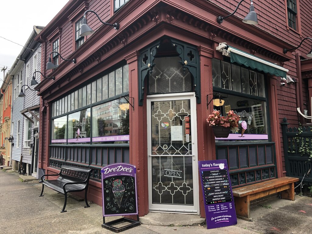 Corner view of Dee Dee’s Ice Cream shop in Halifax, a deep red building with green trim and large windows. A-frame chalkboard outside shows an illustrated ice cream cone, while a purple menu board lists the day’s flavors, including Chocolate, Lemon Blueberry Swirl, and Mint Chocolate Chip. Hanging flowers and a vintage-style glass door marked "OPEN" add to the charm.