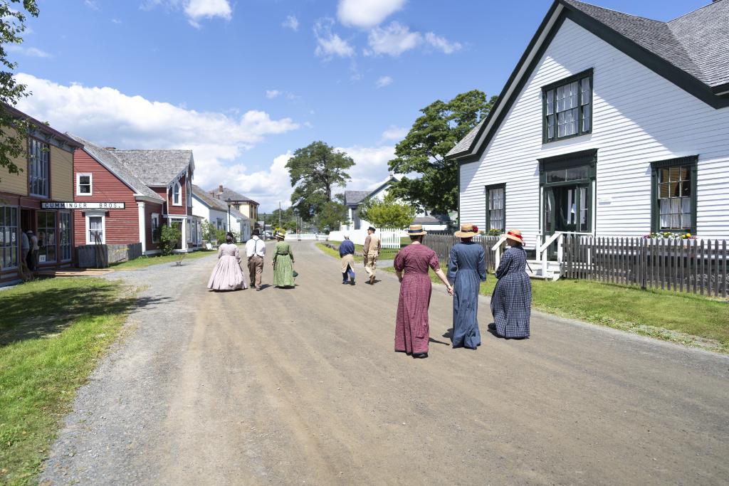 People in 19th-century period clothing walk along a dirt road in Sherbrooke Village, a historical reenactment site on Nova Scotia’s Eastern Shore. Heritage buildings line both sides of the street, including a red storefront marked “Cumminger Bros” and a large white house with green trim.