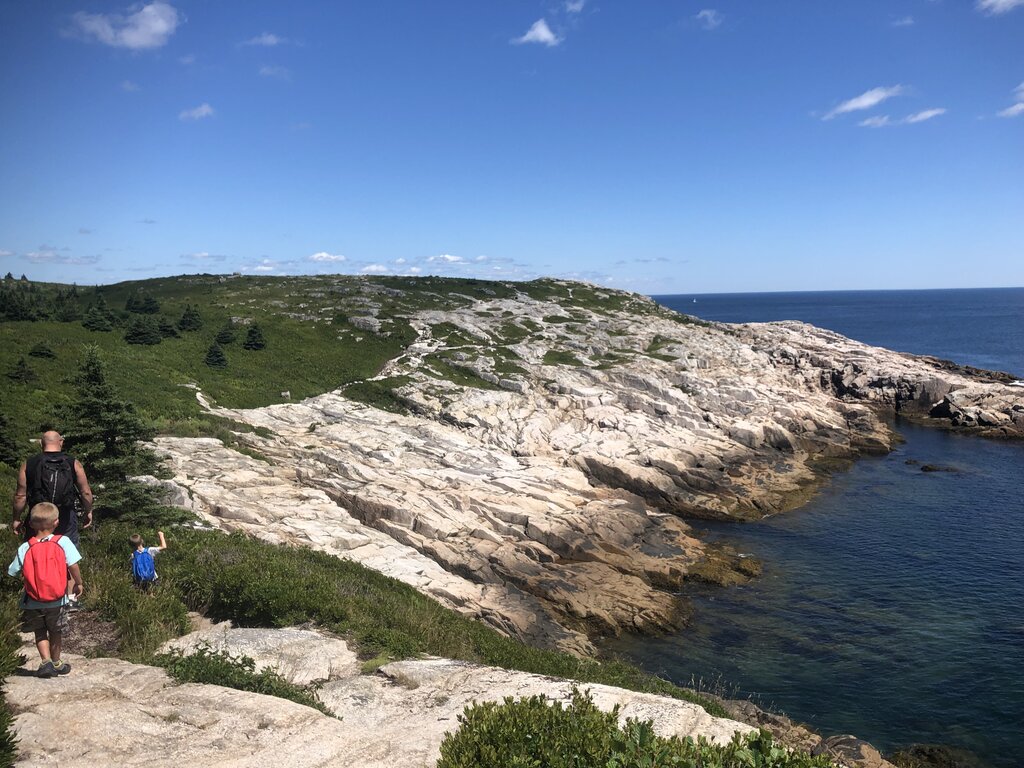 Hikers, including two children with backpacks, walk along a coastal trail at Duncan’s Cove under a clear blue sky. The landscape features smooth, pale granite outcrops meeting deep blue water, with rolling green barrens and scattered evergreens stretching inland.