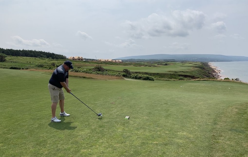 A man tees off on a green golf course overlooking the ocean, with a row of buildings in the distance and rolling hills beyond. The dramatic coastal cliffs and calm blue water highlight the scenic course.