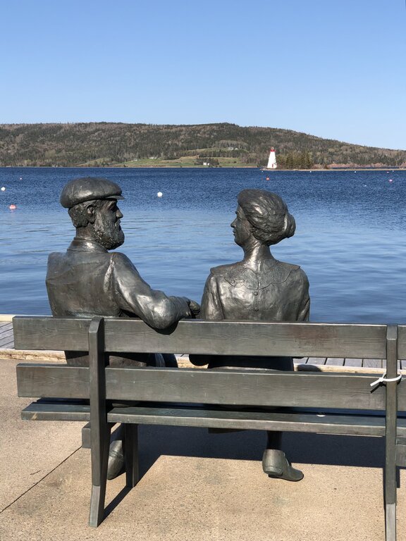 Bronze statues of a man and woman seated on a bench by the waterfront in Baddeck, Nova Scotia, with a lighthouse and hilly shoreline across the water.