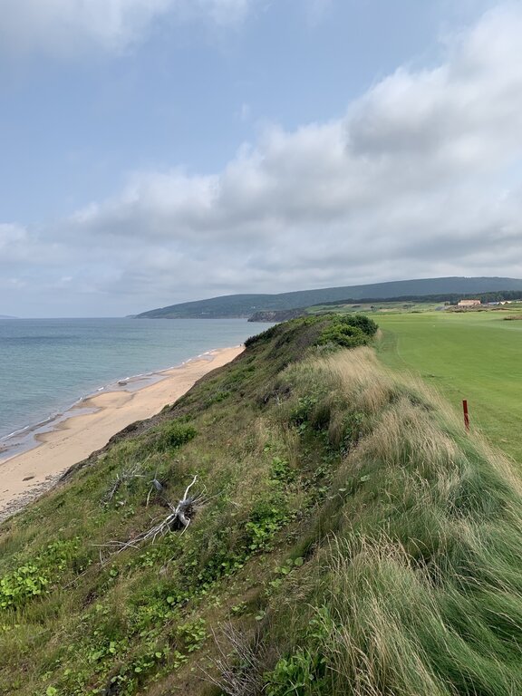 A dramatic coastal view of grassy cliffs dropping to a sandy beach along the edge of a golf course. Ocean waves lap against the shore, and a clubhouse sits in the distance under a lightly clouded sky.