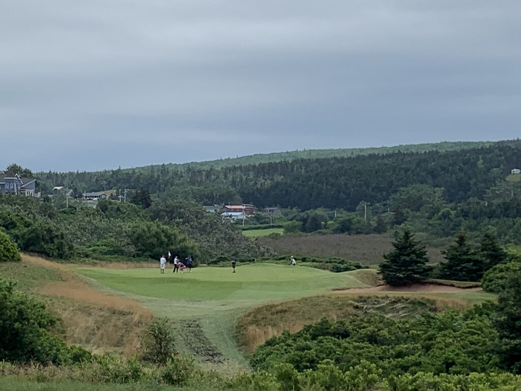 Golfers walk and play on an expansive green course under a cloudy sky, with ocean views on the right and a misty hillside dotted with homes in the distance.