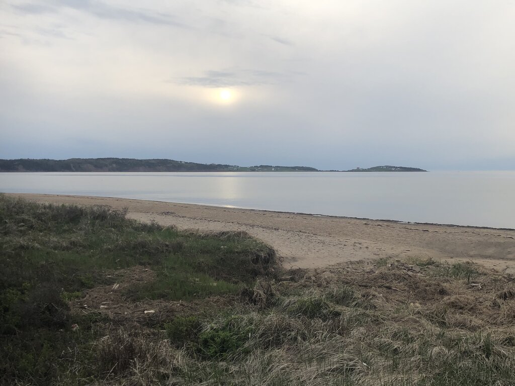 Tranquil beach at Port Hood in Cape Breton with soft evening light, calm water, and low hills across the horizon under a cloudy sky.