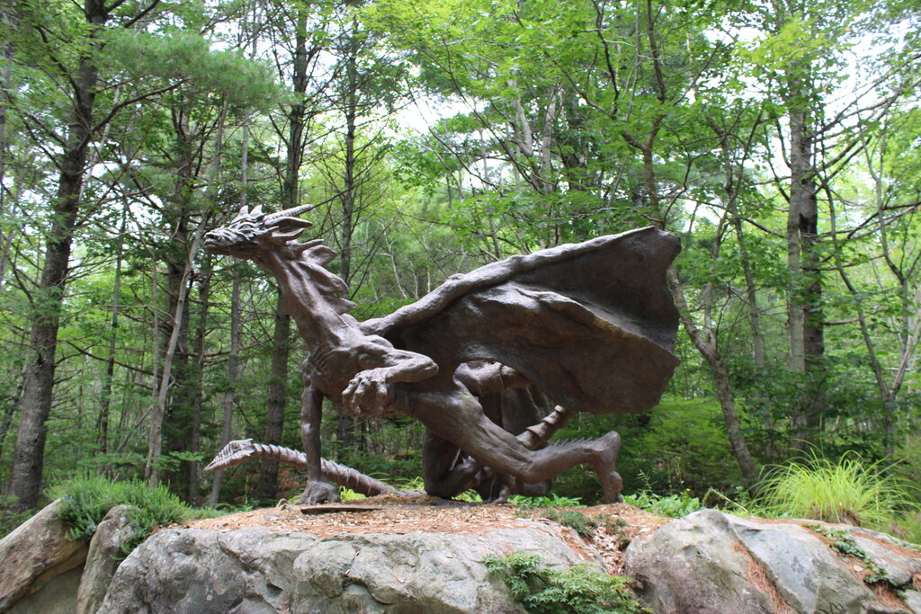 A large dragon sculpture with outstretched wings and sharp features crouches atop a rock platform at Cosby’s Concrete Creations in Liverpool, Nova Scotia. The detailed statue is surrounded by dense forest, creating a mythical atmosphere in the outdoor art garden.