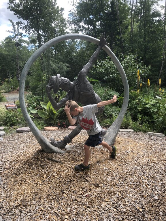 A child playfully mimics the pose of a dynamic sculpture at Cosby’s Concrete Creations garden in Liverpool, Nova Scotia. The statue features a figure frozen mid-leap inside a large circular frame, set among lush greenery and woodchip ground cover.