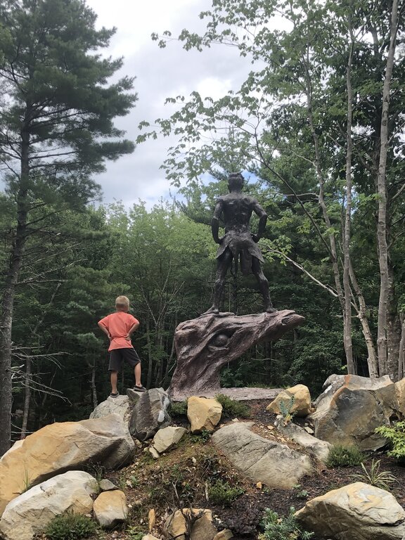 A young boy in a coral t-shirt poses on rocks mirroring a dramatic bronze statue at Cosby’s Concrete Creations garden in Liverpool, Nova Scotia. The statue stands on a twisting pedestal amid large boulders, with tall trees surrounding the outdoor sculpture garden.