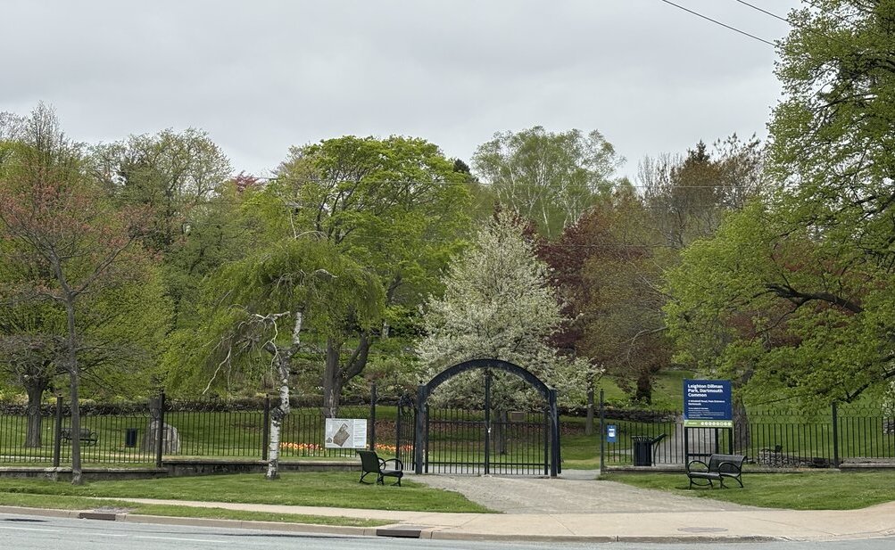 Entrance to Leighton Dillman Park in Dartmouth Commons, marked by a black metal archway and surrounded by colourful spring trees in various shades of green, red, and white.