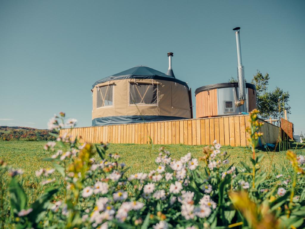 A circular canvas yurt and matching wood-fired hot tub sit elevated on a wooden deck, surrounded by green grass and wildflowers under a clear blue sky. The cozy glamping setup on Nova Scotia’s Eastern Shore offers rustic charm with modern touches, set against a backdrop of rolling hills.
