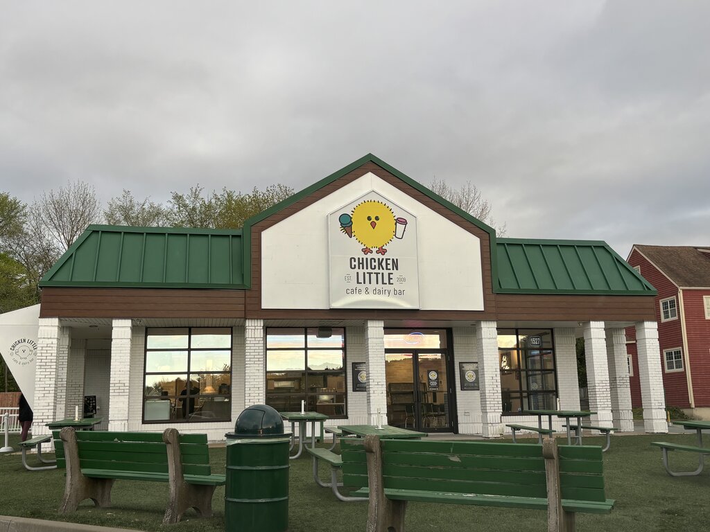 A front view of Chicken Little Café & Dairy Bar, a white brick building with a green metal roof and a large sign featuring a cartoon chick holding an ice cream cone and milkshake. Green picnic benches and a trash bin sit outside on artificial grass under an overcast sky.