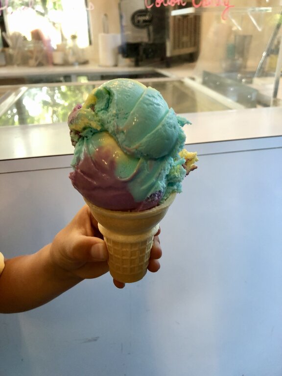 A child holds a colourful scoop of moon mist ice cream in a cone, featuring swirls of blue, purple, and yellow. The background shows an indoor ice cream counter at sugah, one of the best spots for ice cream in Halifax