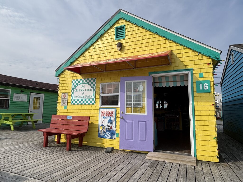 A cheerful yellow wooden building with a teal trim and a purple door, home to Jo’s Old Time Candy & Ice Cream Parlor. A red bench and Slush Puppie sign sit outside, with a boardwalk and other colorful buildings nearby.