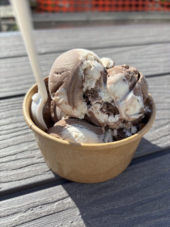 A small brown paper cup filled with scoops of chocolate and vanilla swirled ice cream sits on a sunlit picnic table, with a plastic spoon sticking out.
