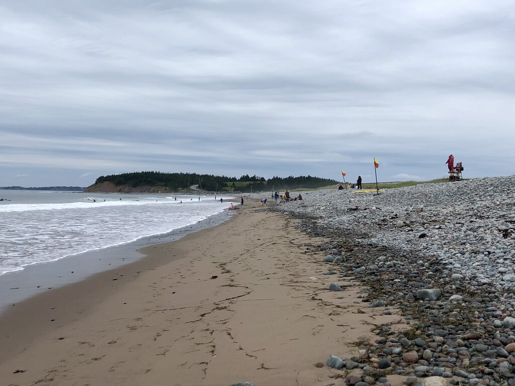 A wide, pebbly beach stretches under a cloudy sky, with a sandy shoreline where people wade into gentle surf. Red and yellow flags mark the lifeguarded area, and a lifeguard in red sits elevated near the rocks.
