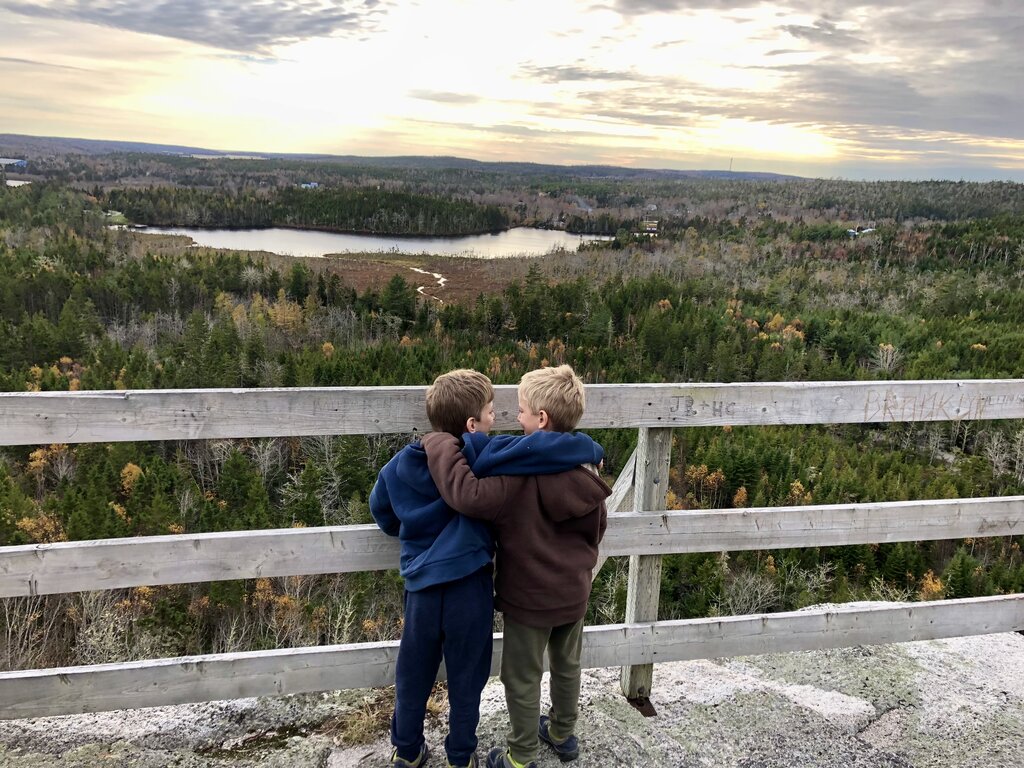 Two young boys stand arm in arm on a lookout platform, gazing over a vast forested valley with a winding river or wetland below, under a softly glowing, cloud-streaked sky at sunset. The wooden railing is carved with initials and overlooks a stunning panoramic view of autumn-toned trees along the Musquodoboit Trail in Nova Scotia.