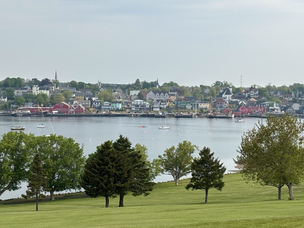 A wide view of Lunenburg, Nova Scotia, shows a colorful waterfront lined with red, yellow, and pastel buildings backed by a hillside of historic homes and church spires, with sailboats anchored in the calm harbor and green trees in the foreground.
