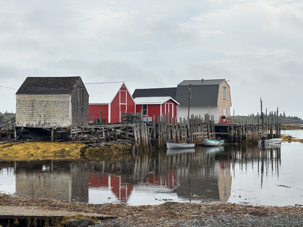 A cluster of weathered and brightly painted fishing shacks on stilts line a wharf in Blue Rocks Nova Scotia, with small boats moored in still water that perfectly reflects the red and grey buildings above.
