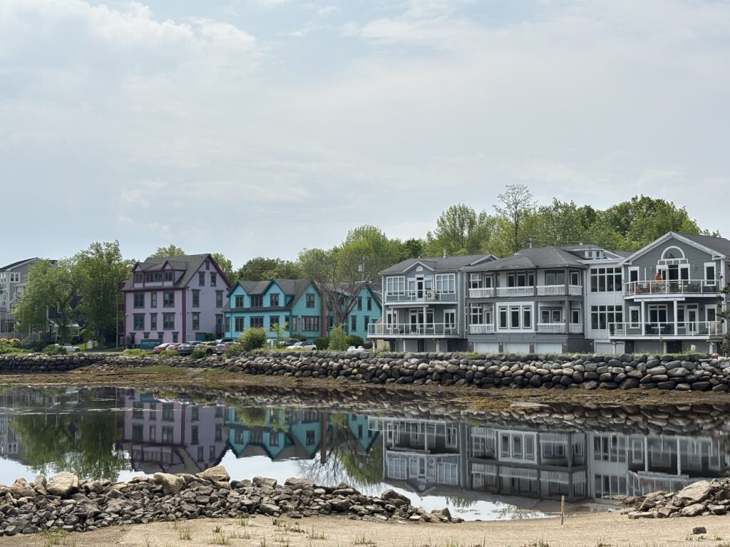 Vibrant purple and turquoise heritage-style houses in Mahone Bay sit beside sleek grey condos along a rocky shoreline, all mirrored crisply in the calm water below on a cloudy day.
