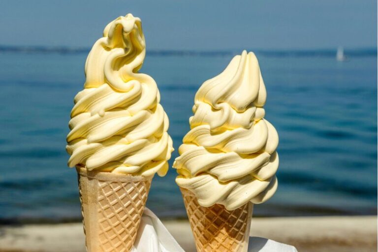 Two tall vanilla soft serve cones in waffle cones held up against a backdrop of calm blue ocean and clear sky. The swirl detail is creamy and perfect, with the seaside view suggesting a relaxed summer day near the Halifax waterfront.
