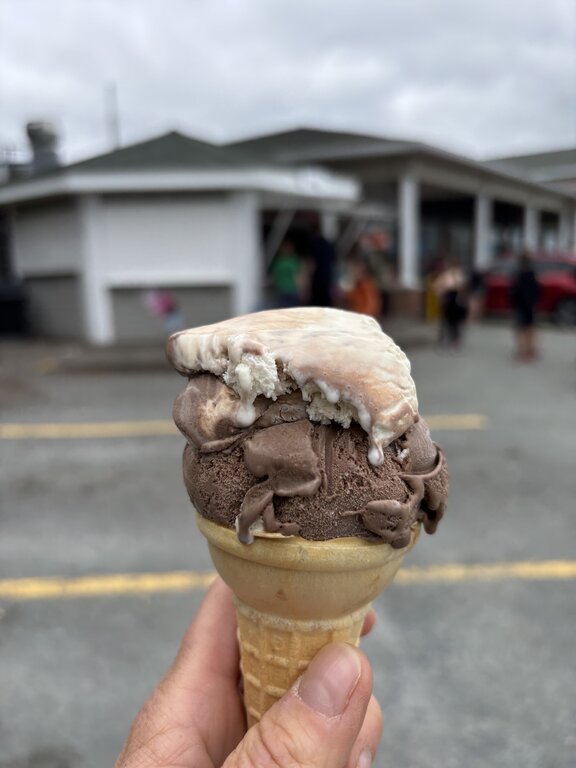 Close-up of a hand holding a waffle cone with two scoops of ice cream—chocolate on the bottom and a partially melted, bite-topped vanilla-chocolate swirl on top. The blurred background shows a small, white building with a lineup of people, likely Colby Scoops, under a grey, overcast sky.