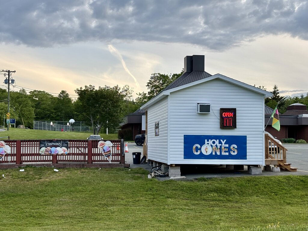 A white ice cream shack called Holy Cones with a large blue sign and a glowing red "OPEN" sign in the window. The small building sits on cinder blocks with a grassy yard out front, and a Chapman’s ice cream banner decorates the wooden fence nearby.