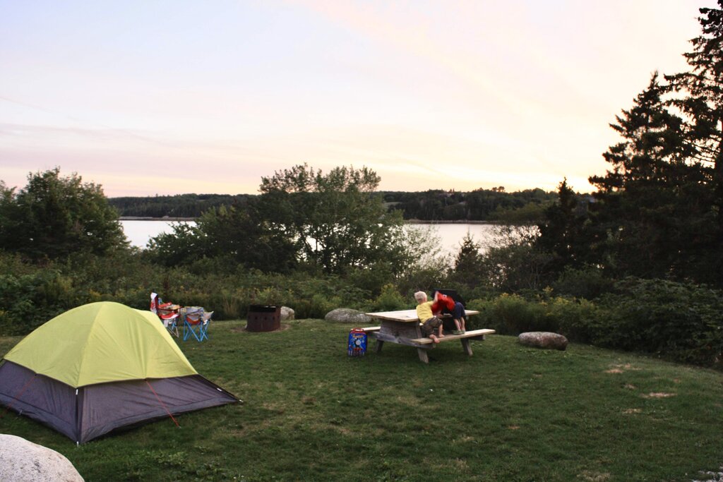 A yellow-and-grey tent pitched lakeside on Graves Island, Nova Scotia, with two children at a picnic table and lush trees backdropping the glowing pink-and-orange sunset over the water.
