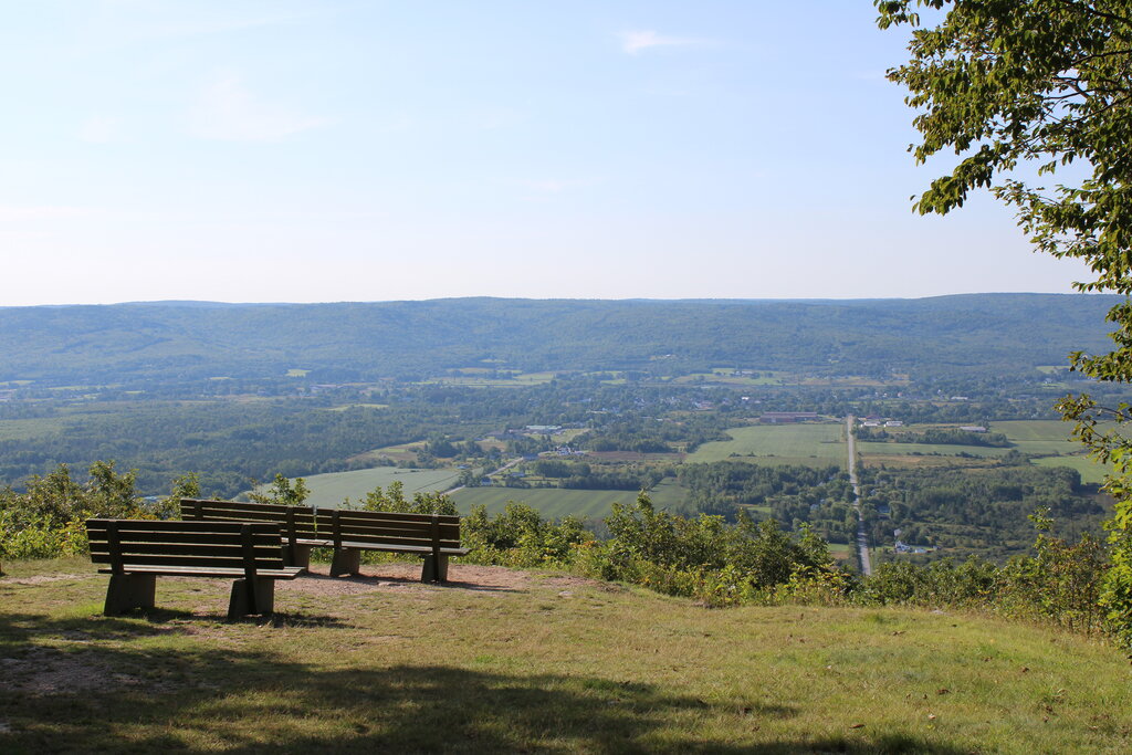 Wooden benches sit on a grassy ridge of a campground overlooking a sweeping view of the Annapolis Valley, with patchwork fields, a straight road, and rolling hills under a clear blue sky.