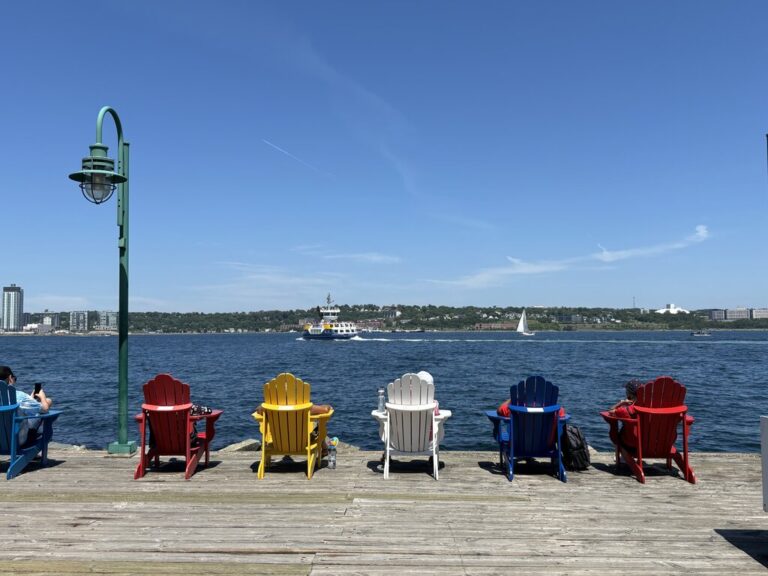 A row of brightly painted Adirondack chairs faces Halifax Harbour from a wooden boardwalk, with a ferry and sailboat visible on the water under a cloudless summer sky.