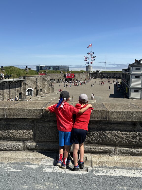 Two children dressed in red shirts embrace as they overlook the bustling main courtyard of Halifax Citadel from a stone wall, with flagpoles and historic buildings in the distance.