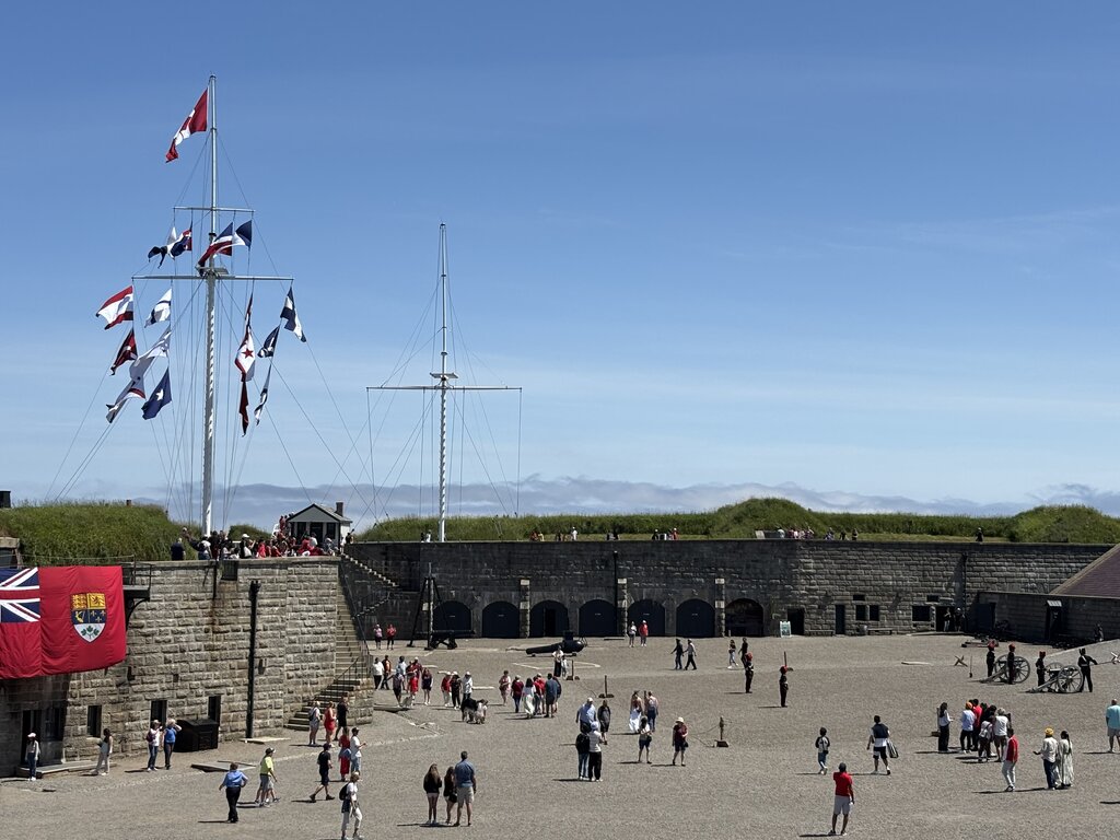 Wide view of Halifax Citadel’s central courtyard shows scattered visitors, costumed guards, and a tall flagpole flying multiple naval and Canadian flags under a clear sky.