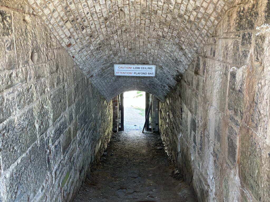 A narrow, arched stone tunnel with a rough brick ceiling leads out of Halifax Citadel, marked with a bilingual sign warning “Caution! Low Ceiling / Attention! Plafond Bas.”