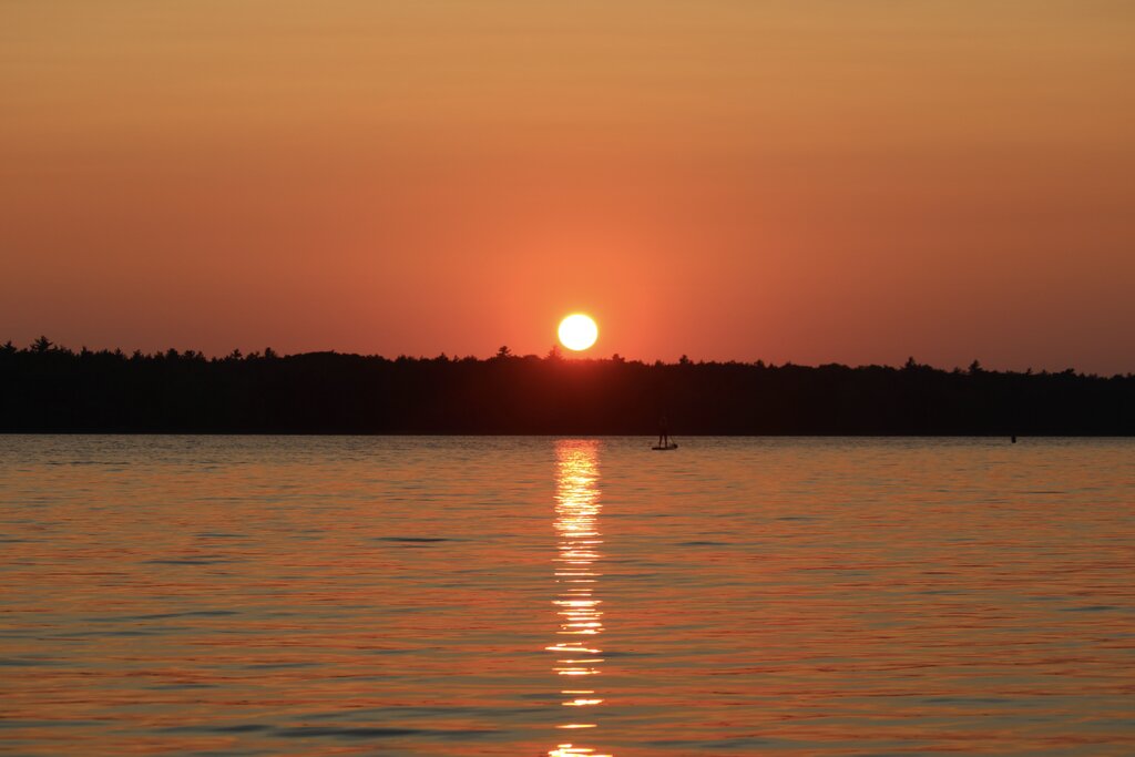 The sun sets over a calm Kejimkujik lake in Nova Scotia, painting the sky orange and reflecting a golden pathway across the water toward a distant dark treeline.