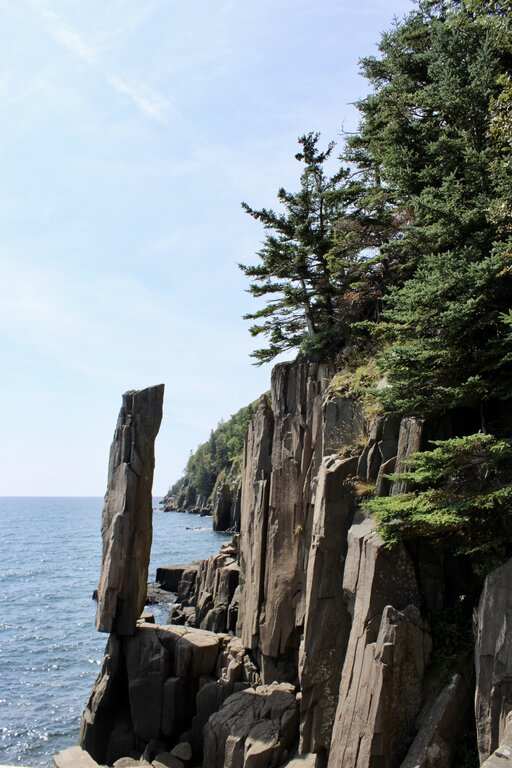 The dramatic Balancing Rock near Digby Neck rises vertically from a jagged basalt cliff, with evergreen trees lining the edge above the calm ocean.