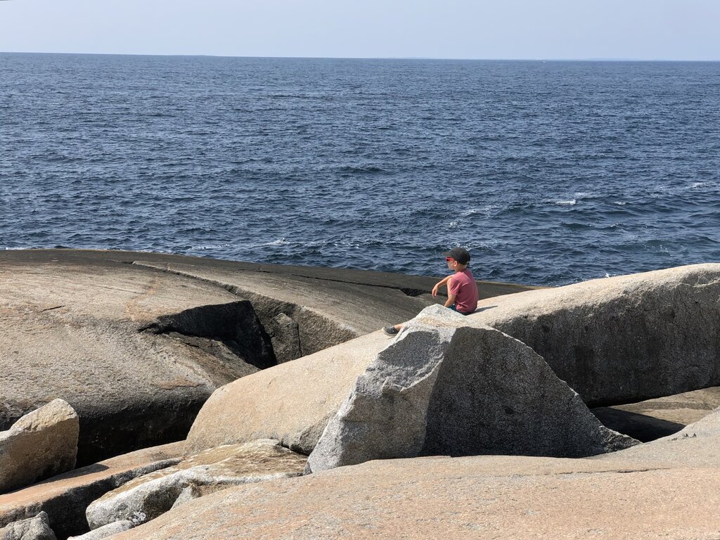 A young boy sits alone on the smooth, weathered granite at Peggy’s Cove, gazing out over the vast Atlantic Ocean under a hazy summer sky.