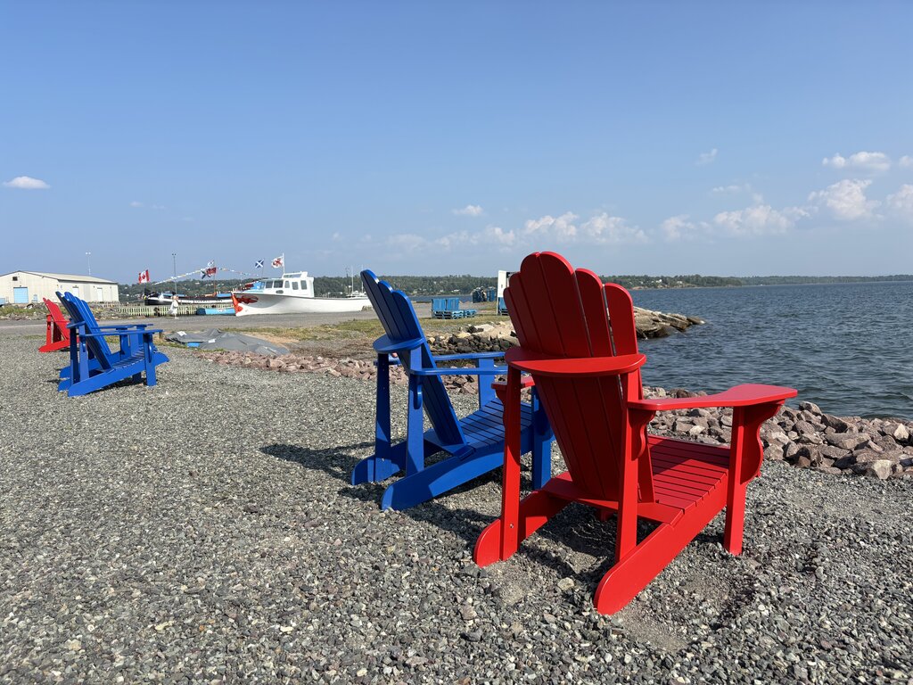 Red and blue Adirondack chairs face the water at the Pictou waterfront, offering a relaxing view of the harbour under a clear blue sky.