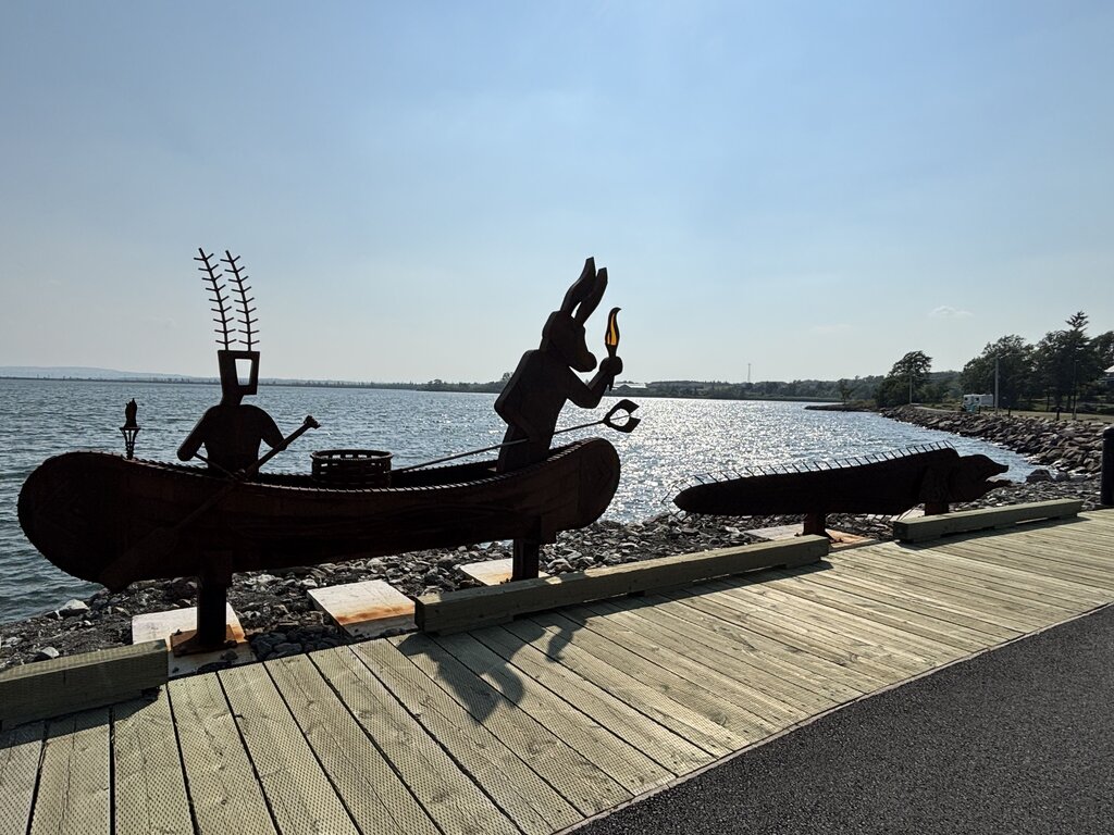 Sculptures of Mi’kmaq figures in a canoe face the water along the Pictou waterfront boardwalk, with the sun glinting off the harbour.
