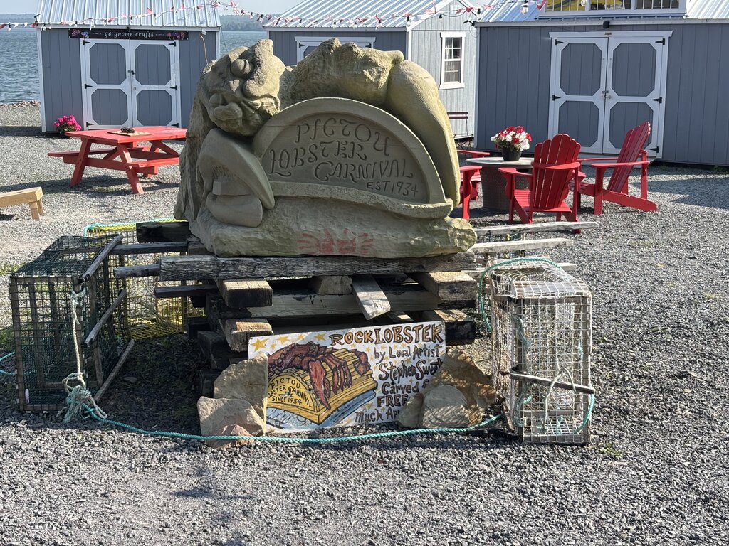 A large carved stone sculpture labeled “Pictou Lobster Carnival EST 1934” sits between lobster traps and handmade signs in front of small craft shops and red chairs.