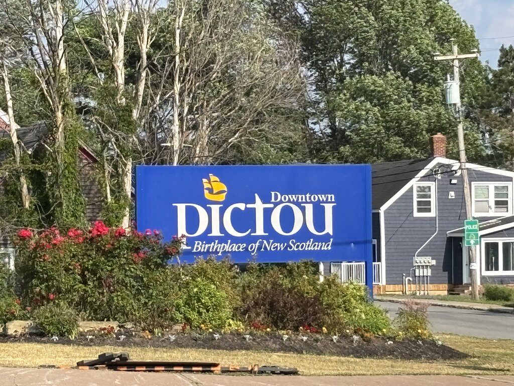 A blue roadside sign reads “Downtown Pictou – Birthplace of New Scotland” set among trimmed bushes and surrounded by homes and tall trees.