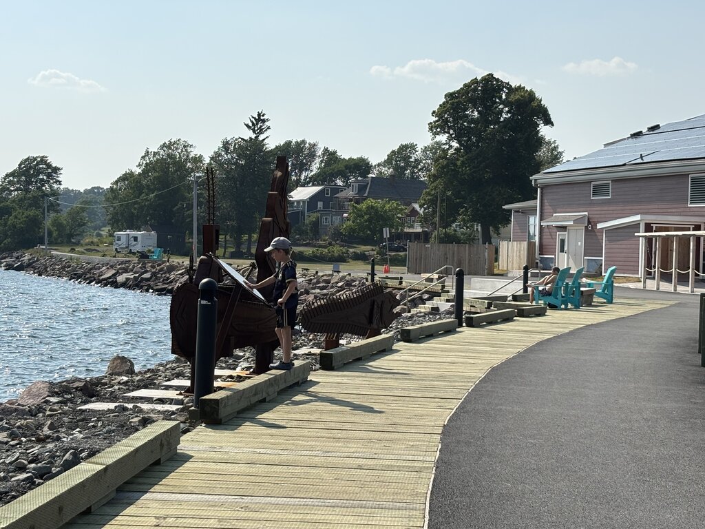 A child stands next to a sculpture of a wooden ship along the Pictou waterfront boardwalk, overlooking the water on a sunny day.