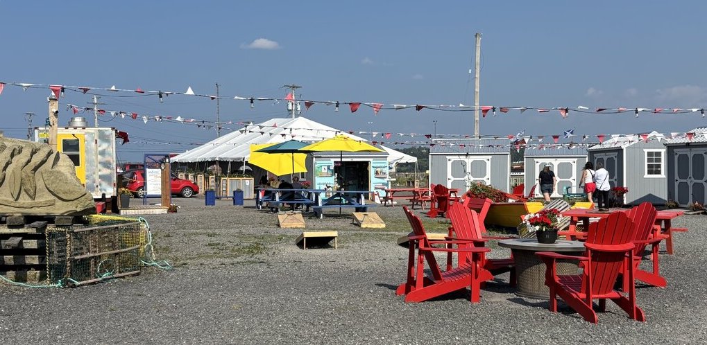 The outdoor market area in Pictou features red Adirondack chairs, bunting flags, food trucks, and local craft huts under a bright summer sky.