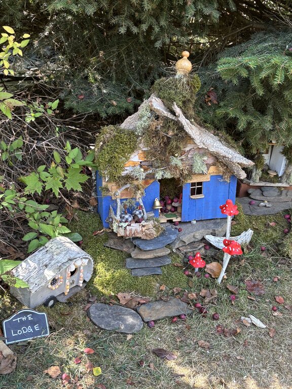 A bright blue fairy house with a moss-covered roof sits beneath evergreen branches, surrounded by red toadstool decorations and a tiny birch log camper marked “The Lodge.”